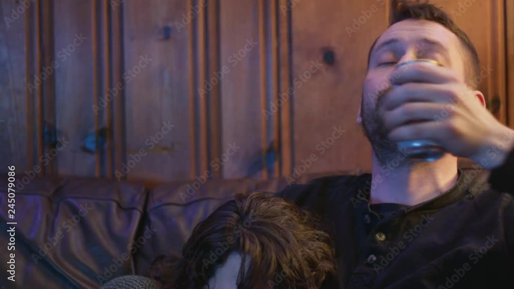 20 something man drinks beer on couch as his cuddled up wife falls asleep below 20 something man drinks beer on couch as his cuddled up wife falls asleep below