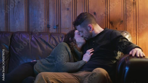 A young couple kiss as they cuddle and snuggle at night on couch, watching tv