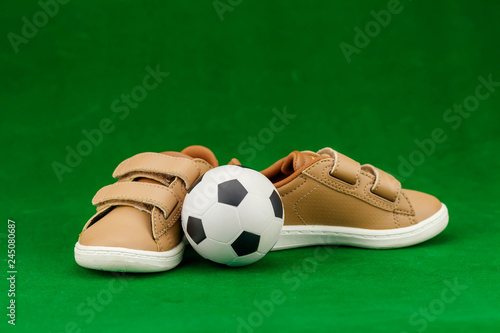 Concept encourage children to play sport  exercise for a healthy body  shoes of small baby shoes next to ball isolated on green background.