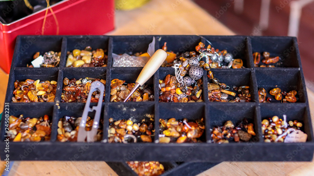 Closeup view at a black tray for handcrafting with multiple sections filled with various kinds of amber, tweezers, stitching awl, needles and a thread