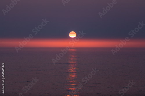 Sunset mirage. Sun  with red  orange and yellow stripes over it. Horizon highlighted in red with a perfect sun path on an ocean surface
