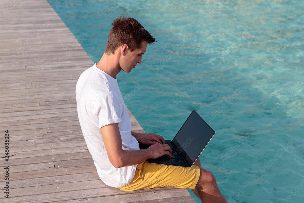 young man seated on a pier and working with his laptop. Clear blue tropical water as background