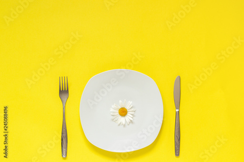One flower chamomile daisy on plate, cutlery fork knife on yellow paper background Concept vegetarian healthy eating diet or anorexia Creative top view Copy space template for lettering text or design