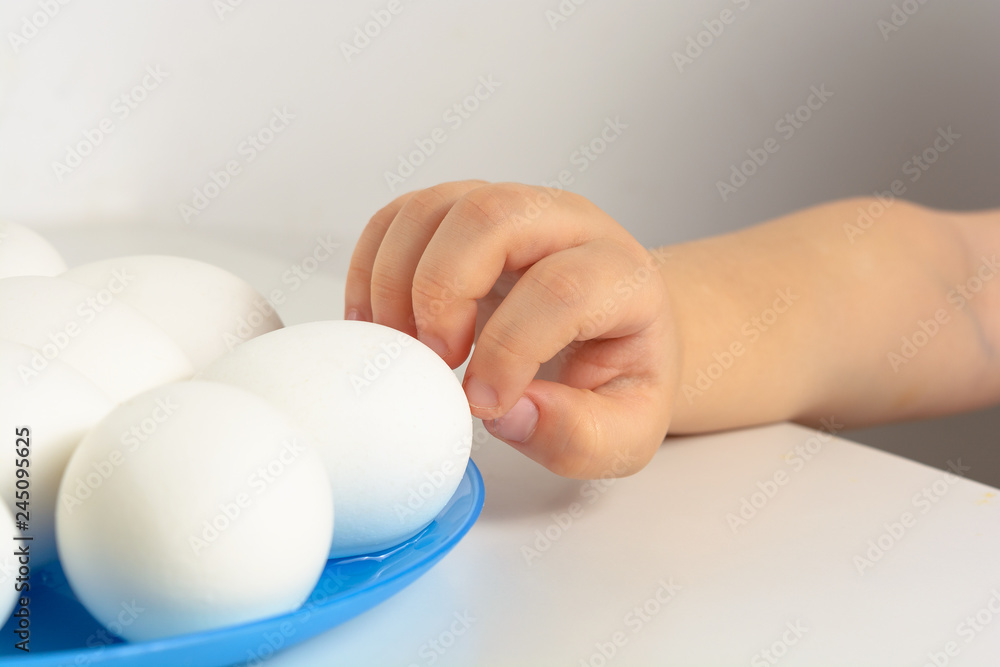 Kid hand stretches for white eggs lying on the blue tlate on the table Kid hand stretches for white eggs lying on the blue tlate on the table