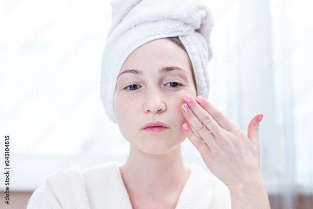 Beautiful happy young woman with a towel on her head looking at her skin in a mirror. Hygiene and care for the skin