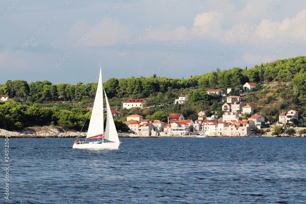 A modern cruise sailing yacht with a Bermuda sloop-type rig goes past the green coast of the Croatian Riviera on a sunny summer day. Adriatic Sea of the Mediterranean region. District of Dalmatia A modern cruise sailing yacht with a Bermuda sloop-type rig goes past the green coast of the Croatian Riviera on a sunny summer day. Adriatic Sea of the Mediterranean region. District of Dalmatia