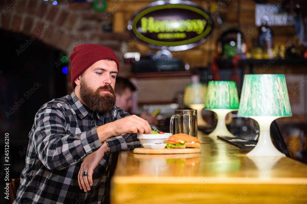 Hipster hungry man eat burger. Man with beard eat burger menu. Brutal hipster bearded man sit at bar counter. Cheat meal. High calorie food. Delicious burger concept. Enjoy taste of fresh burger