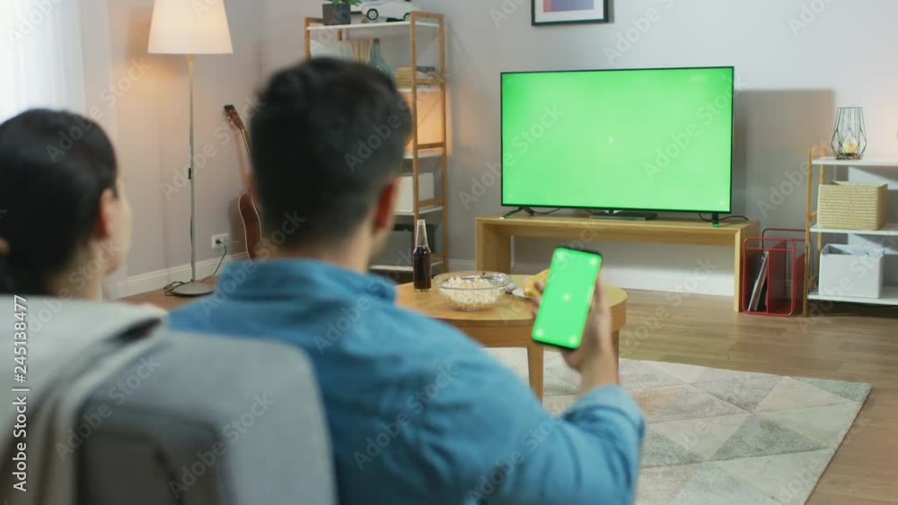 Happy Couple Sitting At Home in the Living Room Watching Green Chroma Key Screen Television, Relaxing on a Couch. Guy also Uses Green Mock-up Screen Smartphone. Happy Couple Sitting At Home in the Living Room Watching Green Chroma Key Screen Television, Relaxing on a Couch. Guy also Uses Green Mock-up Screen Smartphone.
