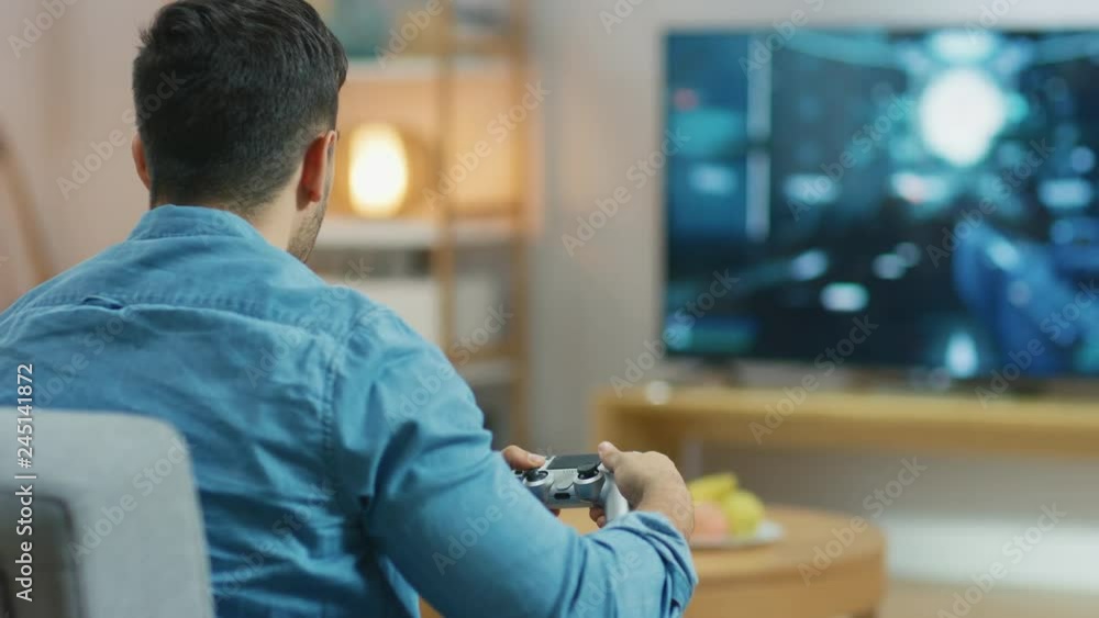 In the Living Room Man Sitting on a Couch Holds Controller Playing in a Console Video Game, 3D Action Shooter Gameplay Shown on TV Screen. In the Living Room Man Sitting on a Couch Holds Controller Playing in a Console Video Game, 3D Action Shooter Gameplay Shown on TV Screen.