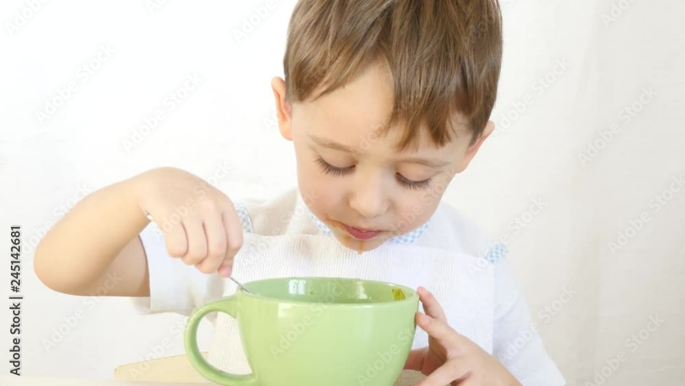 The child with pleasure eat soup from dishes with a spoon, close-up. A boy eats sitting at a table on a white background. The child with pleasure eat soup from dishes with a spoon, close-up. A boy eats sitting at a table on a white background.
