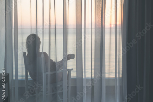 Naked beautiful female wrapped in a blanket sitting on a chair with cup of coffee behind the transparent curtains on the glass balcony early morning at sunrise. Sea view