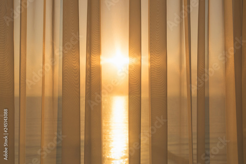 Window of a balcony with transparent curtains and view of morning sun and sea