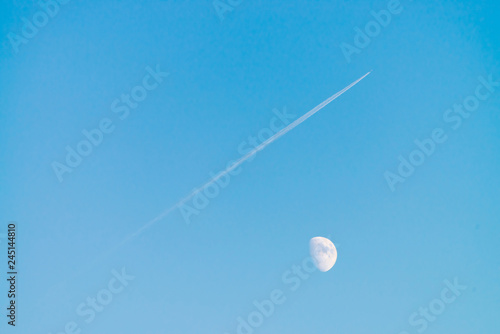 Condensation track of jet above moon in clear blue day sky. Minimalist blue background. Plane flies up diagonally. Airplane is flying in air space. White moon in cyan sky.