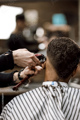 The barber makes a razor cut hair back and sides for a stylish black-haired man sitting in the armchair in a barbershop