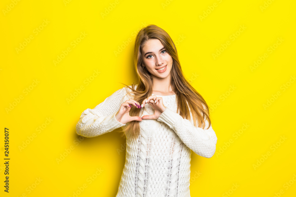 Smiling young girl in white sweater showing heart with two hands, love sign isolated over yellow background. Smiling young girl in white sweater showing heart with two hands, love sign isolated over yellow background.