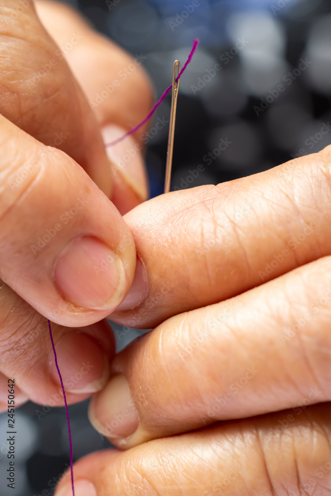 Senior woman's hands finishing to thread a needle, Purple colour roll of thread, Close up & Macro shot, Selective focus, Blurred and bokeh background, Tailor, Needlework concept Senior woman's hands finishing to thread a needle, Purple colour roll of thread, Close up & Macro shot, Selective focus, Blurred and bokeh background, Tailor, Needlework concept