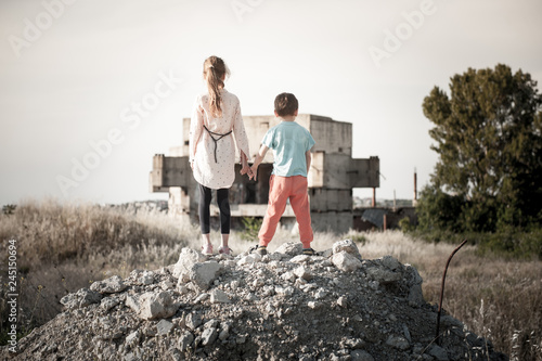 two poor refugees children standing with hope together holding hands on ruins of destroyed during war fighting home house photo