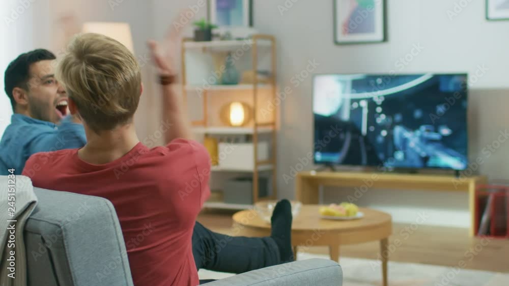 In the Living Room Two Friends Sitting on a Couch Holding Controllers Playing Competitive Video Game, 3D Action Shooter Gameplay Shown on TV Screen. They Do High Five After Finishing Level In the Living Room Two Friends Sitting on a Couch Holding Controllers Playing Competitive Video Game, 3D Action Shooter Gameplay Shown on TV Screen. They Do High Five After Finishing Level