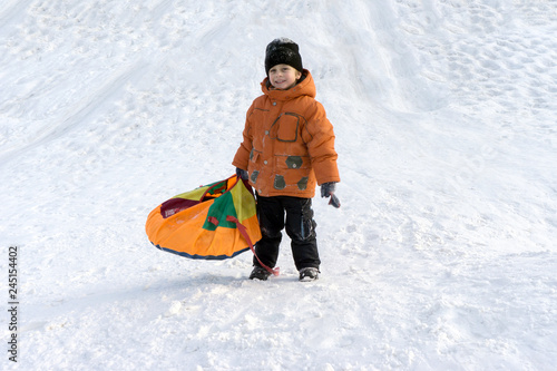 a boy in an orange jacket and a black hat on a Sunny winter day with a snow slide on a special bun and laughs