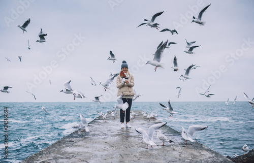 Happy young tourist woman feeds seagulls on the sea. Pretty female wearing coat, scarf and watching flying seagulls by the sea in the sky. People, travel, environment, nature concept.