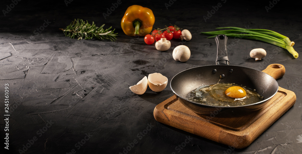 Chicken egg in empty pan surrounded by vegetarian cuisine ingredients on dark table