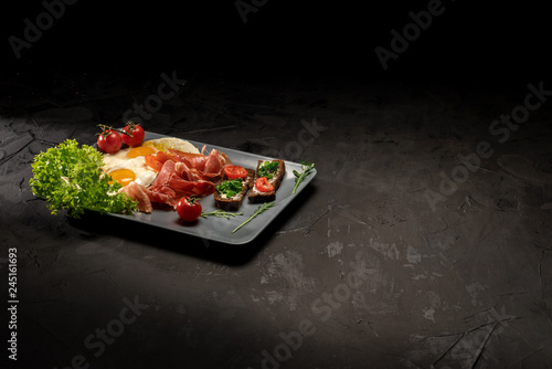 Healthy breakfast with fried eggs, bacon, toasts, tomatoes and fresh salad on black background