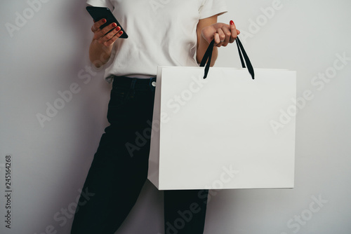 Hipster girl in casual white t-shirt holding smartphone and blank paper bag with copy space for logo or design, mock-up of white shopping package, white wall background, shopping online concept