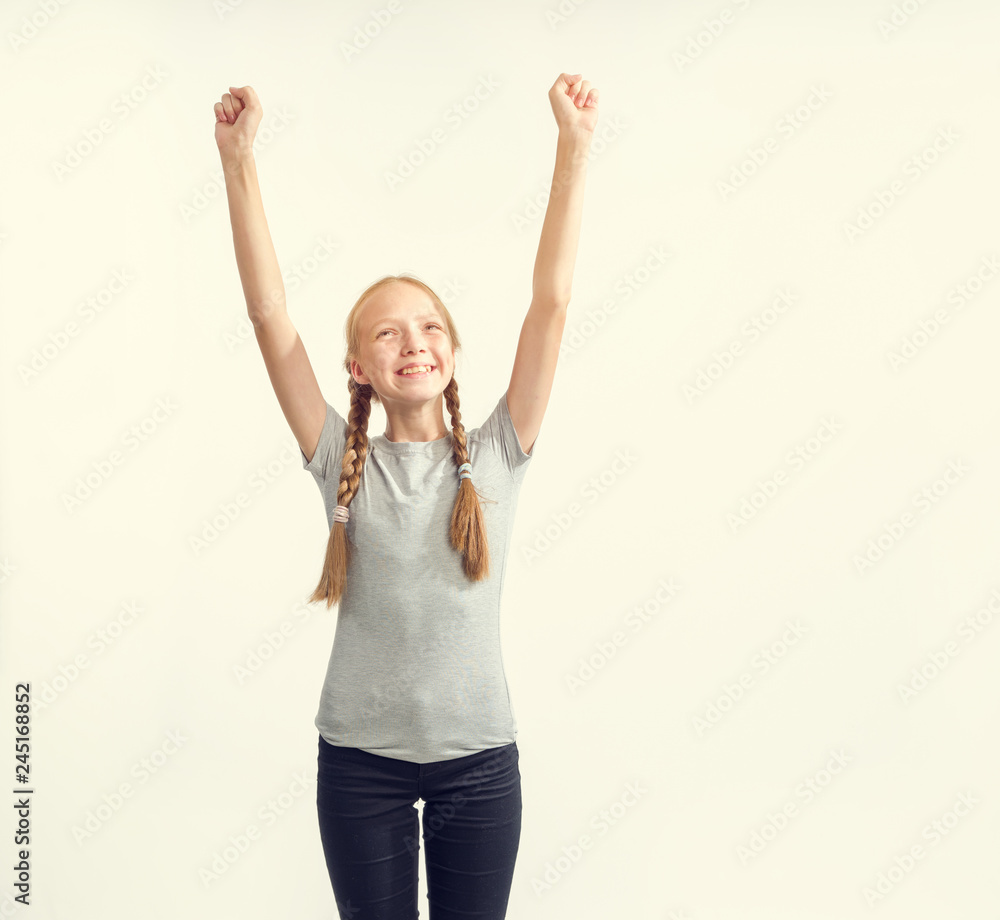 Portrait of a fun 13 years old girl on a light background. The girl in the gray t-shirt raised her hands up. She is joyful.