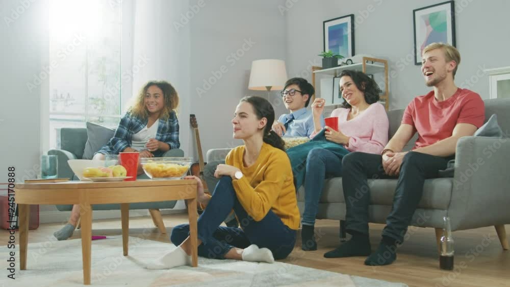 At Home Diverse Group Friends Watching TV Together, Eating Snacks and Drinking Beverage. They Probably Watching Sports Game, Movie or Sitcom TV Show. Young People Having Fun Together. At Home Diverse Group Friends Watching TV Together, Eating Snacks and Drinking Beverage. They Probably Watching Sports Game, Movie or Sitcom TV Show. Young People Having Fun Together.