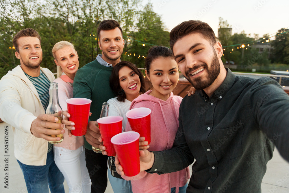 Fototapeta premium leisure and people concept - happy friends with drinks taking selfie at rooftop party in summer