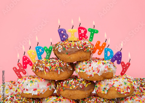 Chocolate and white frosted donuts covered in candy sprinkles piled into a cake pile with Happy Birthday candles burning brightly. Pink background.