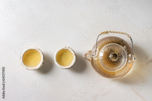 Hot green tea in two traditional chinese clay ceramic cup and glass teapot in row over white marble background. Flat lay, space