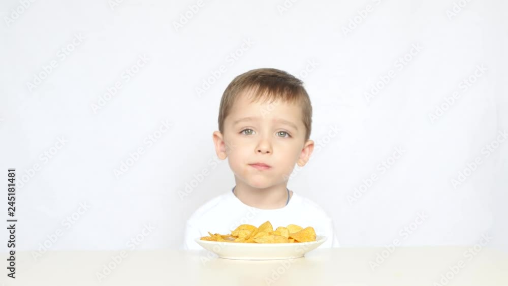 A cute little boy is sitting at the table in front of a plate of chips on a white background. A child eats potato chips with gusto. Not a healthy diet child. A cute little boy is sitting at the table in front of a plate of chips on a white background. A child eats potato chips with gusto. Not a healthy diet child.