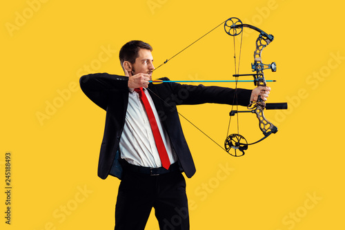 Businessman aiming at target with bow and arrow, isolated on yellow studio background. The business, goal, challenge, competition, achievement concept