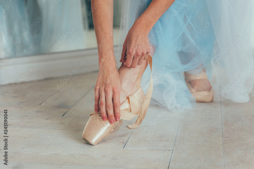 Hands of ballerina in blue tutu skirt puts on pointe shoes on leg in white light hall. Young classical ballet dancer woman in dance class Hands of ballerina in blue tutu skirt puts on pointe shoes on leg in white light hall. Young classical ballet dancer woman in dance class