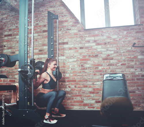 Foto Smilling athletic woman exercising squatting with barbell at smith machine against brick wall in gym