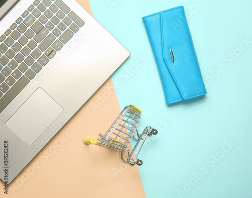Online shopping. Mini shopping trolley, laptop, blue leather purse on a colored pastel background, top view.