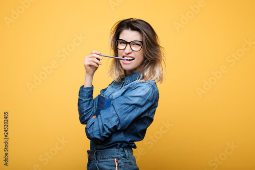 Portrait of a amazing female with eyeglases dressed in blue jeans clothes with dyed hair bitting a pencil and looking into camera against a yellow blackground.  photo