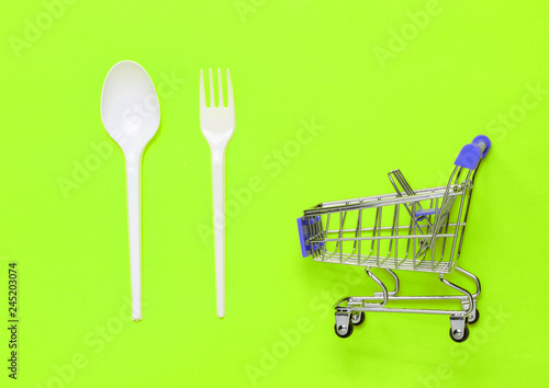 Shopping trolley, plastic spoon and fork on green background. Restaurant food, purchase of food products.