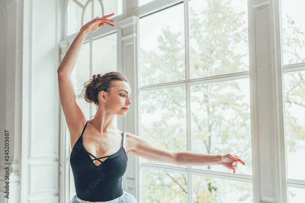 Young classical ballet dancer woman in dance class. Beautiful graceful ballerina practice ballet positions in blue tutu skirt near large window in white light hall