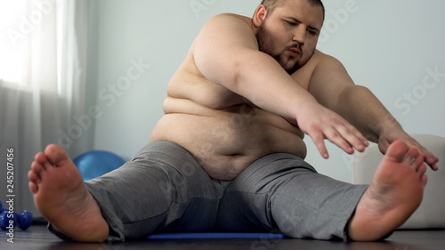 Oversize man stretching on mat trying to reach toes, improving muscle elasticity