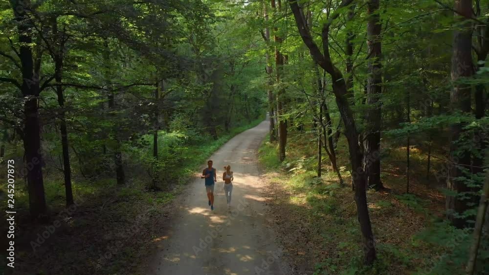 DRONE: Flying in front of a fit woman and man running through the sunny forest. Athletic young couple jogging down an empty forest trail on a sunny summer afternoon. Healthy boyfriend and girlfriend. DRONE: Flying in front of a fit woman and man running through the sunny forest. Athletic young couple jogging down an empty forest trail on a sunny summer afternoon. Healthy boyfriend and girlfriend.