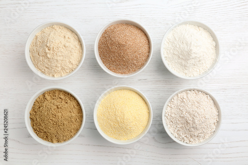 Bowls with different types of flour on white wooden background, top view