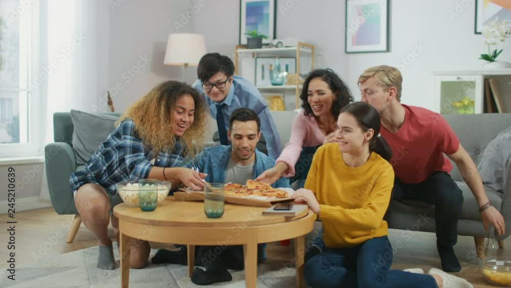 At Home Diverse Group Friends Watching TV Together, Girl Brings Gigantic Pizza and Friends Share it. Guys and Girls Watching Comedy Sitcom or a Movie, Laughing and Having Fun Together. At Home Diverse Group Friends Watching TV Together, Girl Brings Gigantic Pizza and Friends Share it. Guys and Girls Watching Comedy Sitcom or a Movie, Laughing and Having Fun Together.