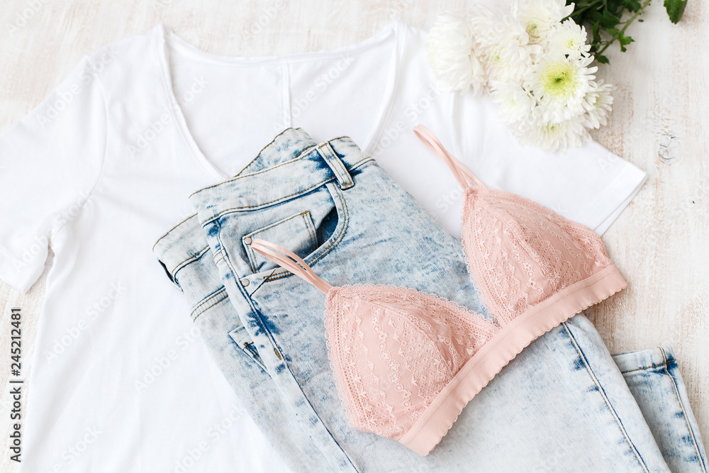 White t-shirt, blue jeans, pink lace triangle bra, white flower on white  wooden background. Overhead