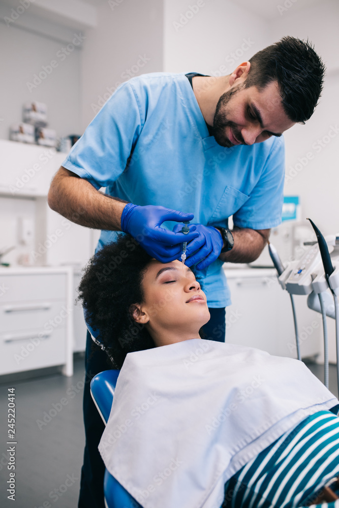 Attractive young African woman is getting a rejuvenating facial injections. She is sitting calmly at clinic. The expert beautician is filling female wrinkles by hyaluronic acid. Attractive young African woman is getting a rejuvenating facial injections. She is sitting calmly at clinic. The expert beautician is filling female wrinkles by hyaluronic acid.