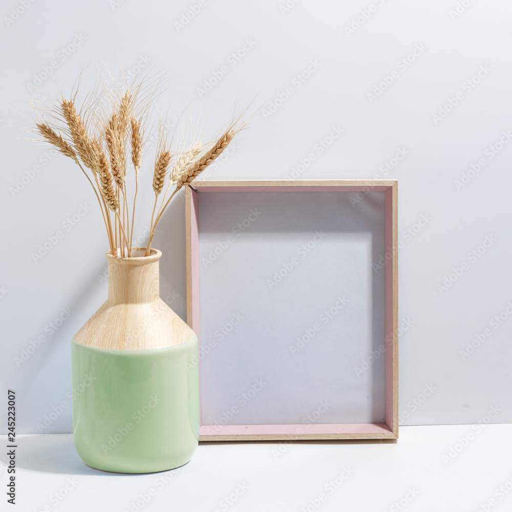 Mock up white frame and spikelets of wheat in ligth-green vase on book shelf or desk. Minimalistic concept.