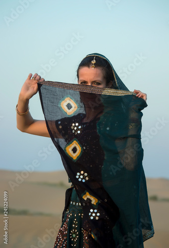 A girl in Indian clothes hides her face behind a veil against the sky and dunes. Rajasthan, India.