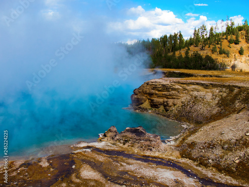 A vibrant blue geothermal hot spring in Yellowstone National Park emits thick steam, creating a mystical landscape surrounded by rugged terrain, lush forest, and a bright blue sky with scattered cloud