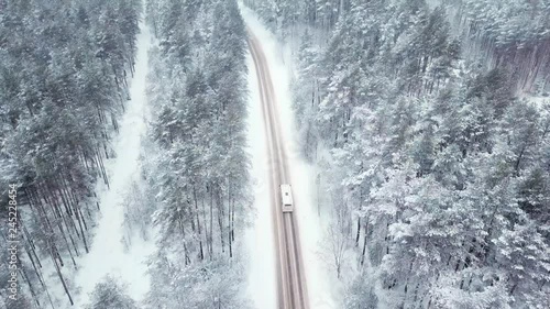 A bus is driving along a snowy road in the middle of a winter frozen forest. Firs and pines in the snow. Snowing. Aerial view